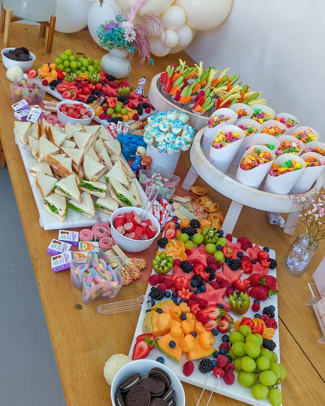 Party food spread with fruit platters, sandwiches, and colorful candies on a wooden table.