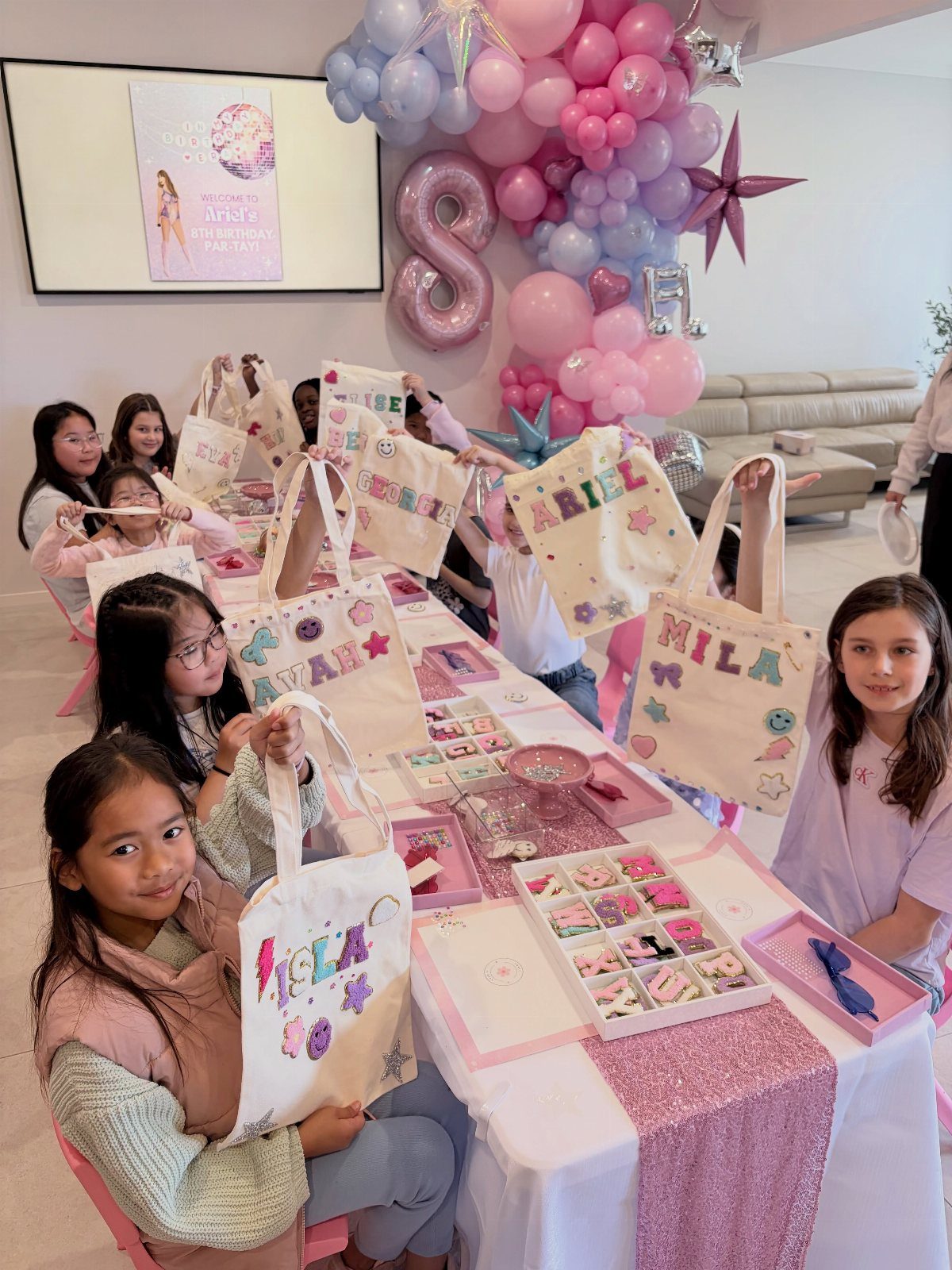 Young girls holding up decorated tote bags at a birthday craft party table.