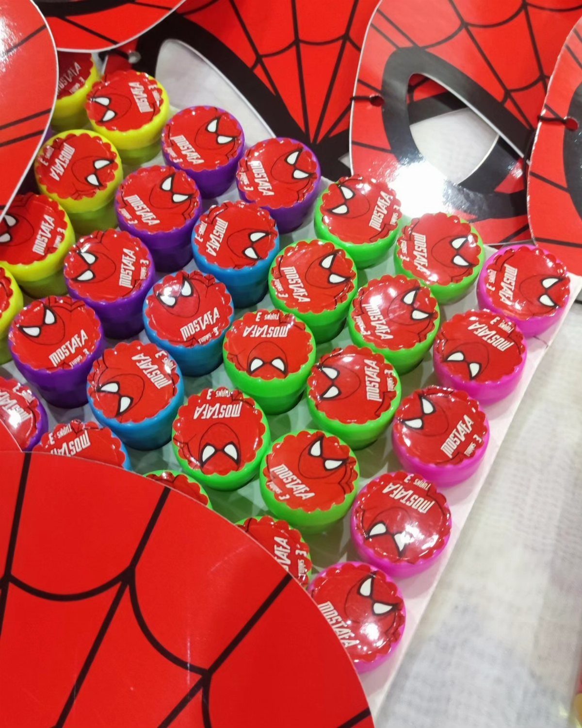 Spider-Man themed party favor containers in various colors with personalized red lids.