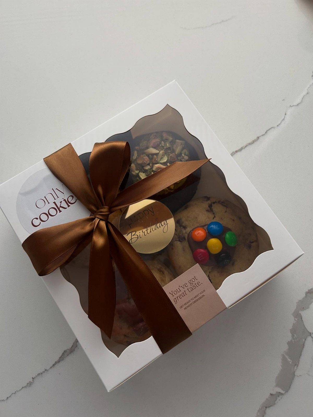 White cookie gift box with a brown bow and colorful chocolate cookies inside.