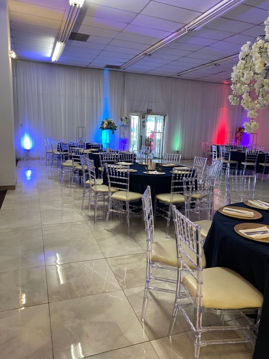 Banquet hall with round tables, black linens, clear chairs, and colorful accent lighting.