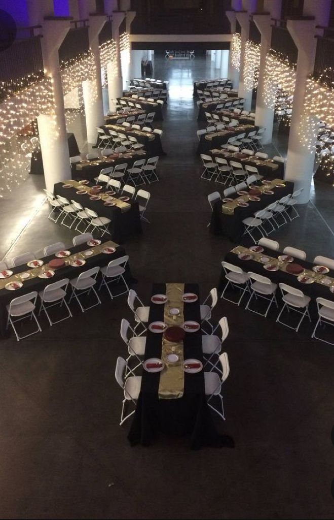 Event hall with long black tables, gold runners, white chairs, and string lights.