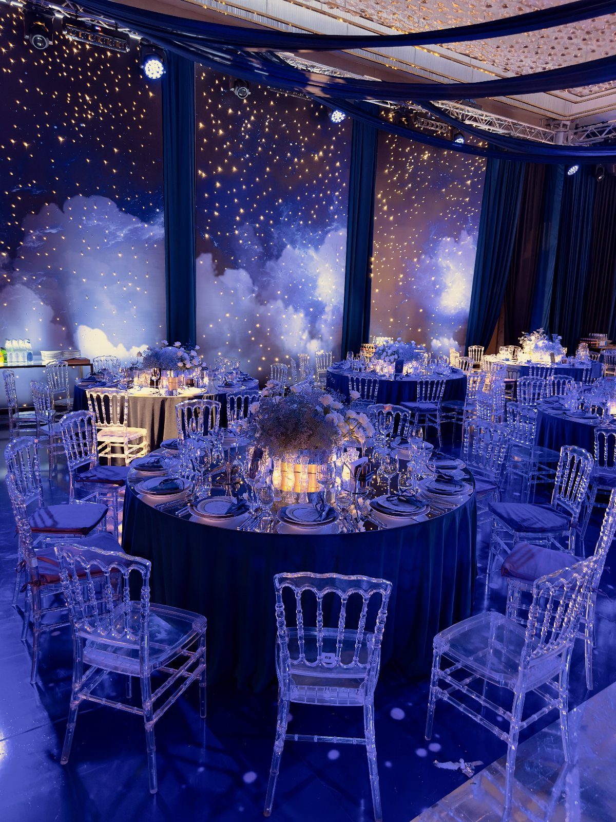 Event hall with round tables, clear chairs, and a starry night sky projection.