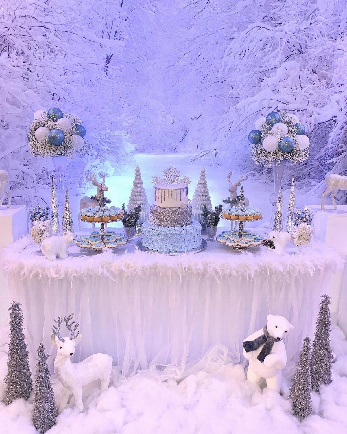 Winter themed dessert table with blue cake, white faux snow, and reindeer figurines.