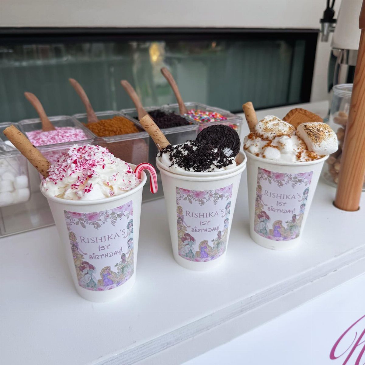 Three decorated hot chocolate cups with whipped cream and toppings at a party station.