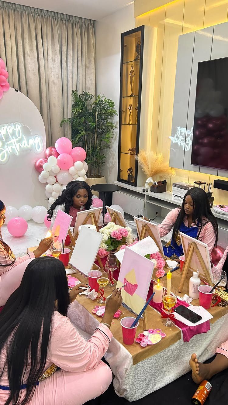 Women in pink painting on canvases at a birthday party with balloon decorations.