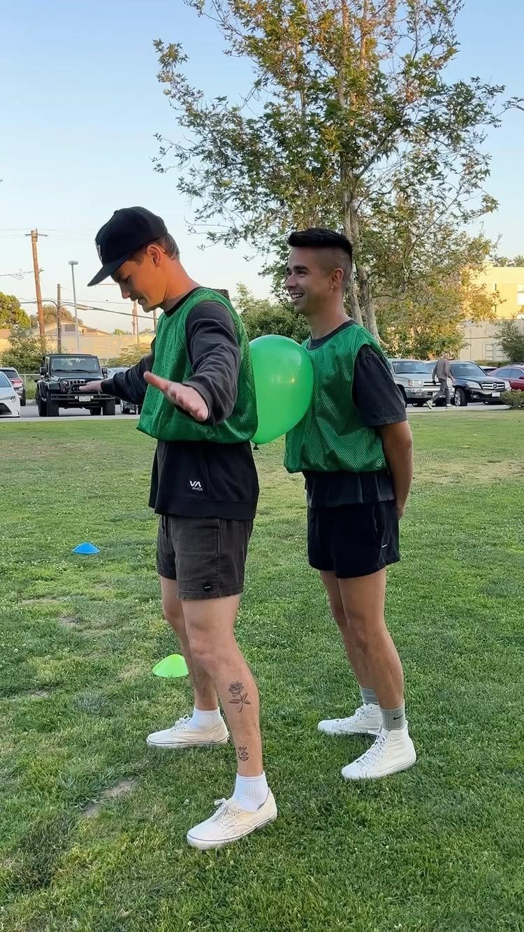 Two men in green vests balancing a green balloon between their backs outdoors.
