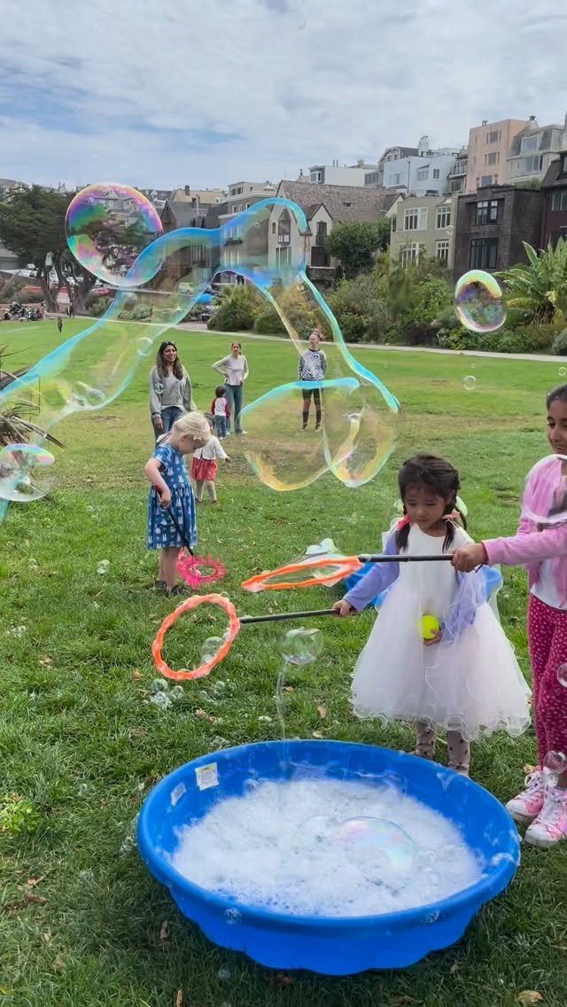 Children making giant iridescent bubbles with wands in a grassy city park.