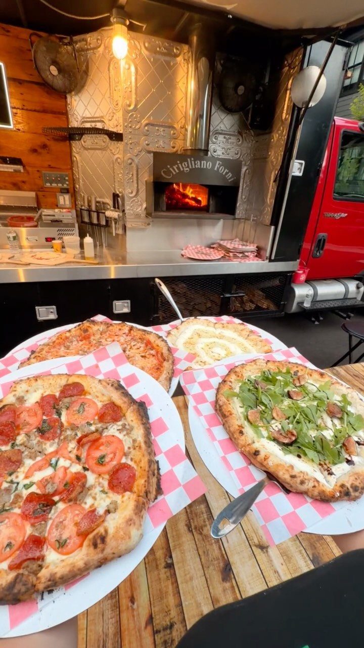 Three fresh pizzas on a wooden table in front of a red food truck.