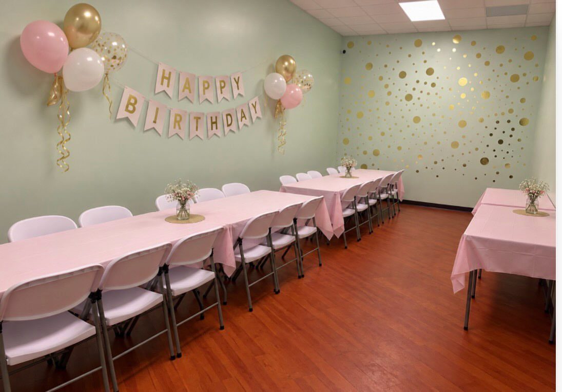 Birthday party room with pink tablecloths, white chairs, and gold balloon decorations.