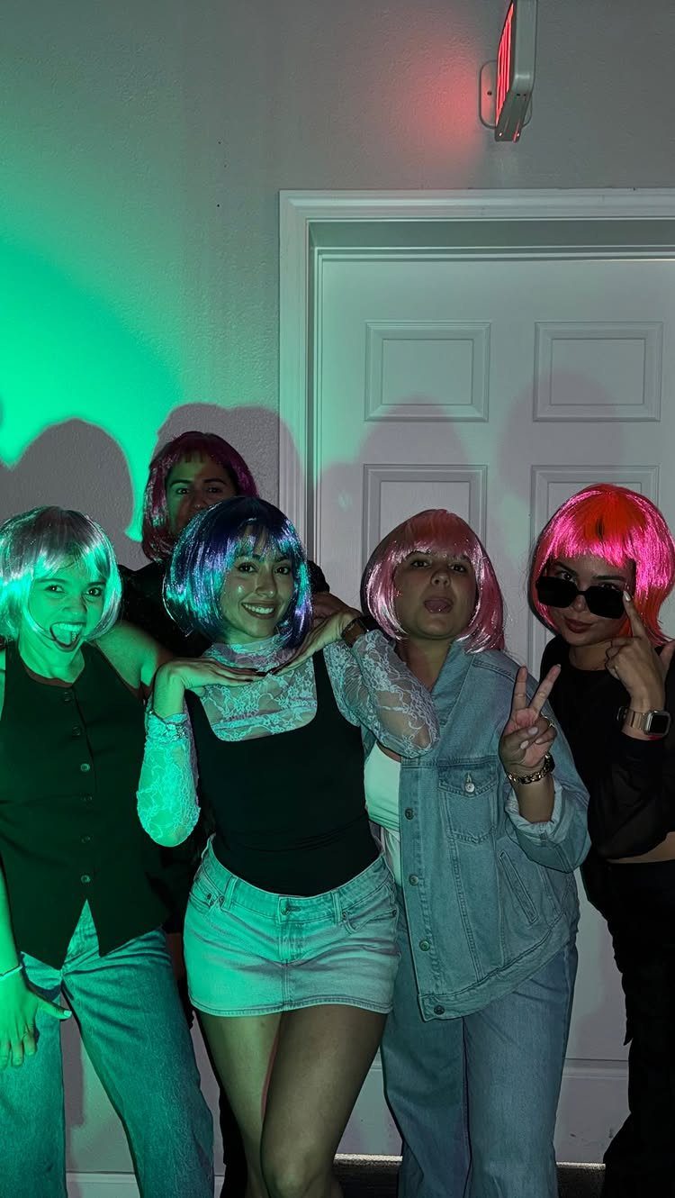 Five young women wearing colorful wigs posing together under green and red lights.