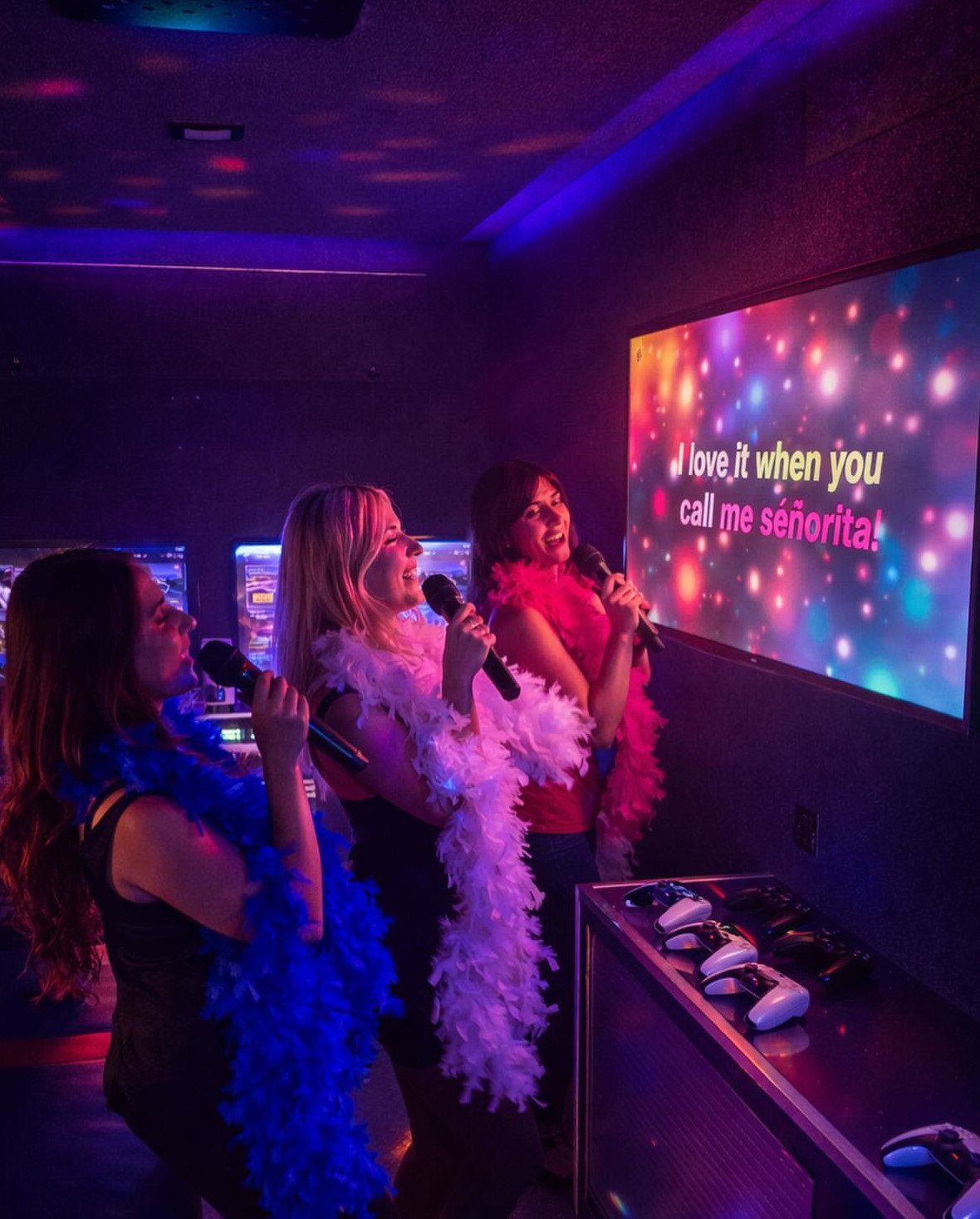 Three women singing into microphones with feather boas during a karaoke party.