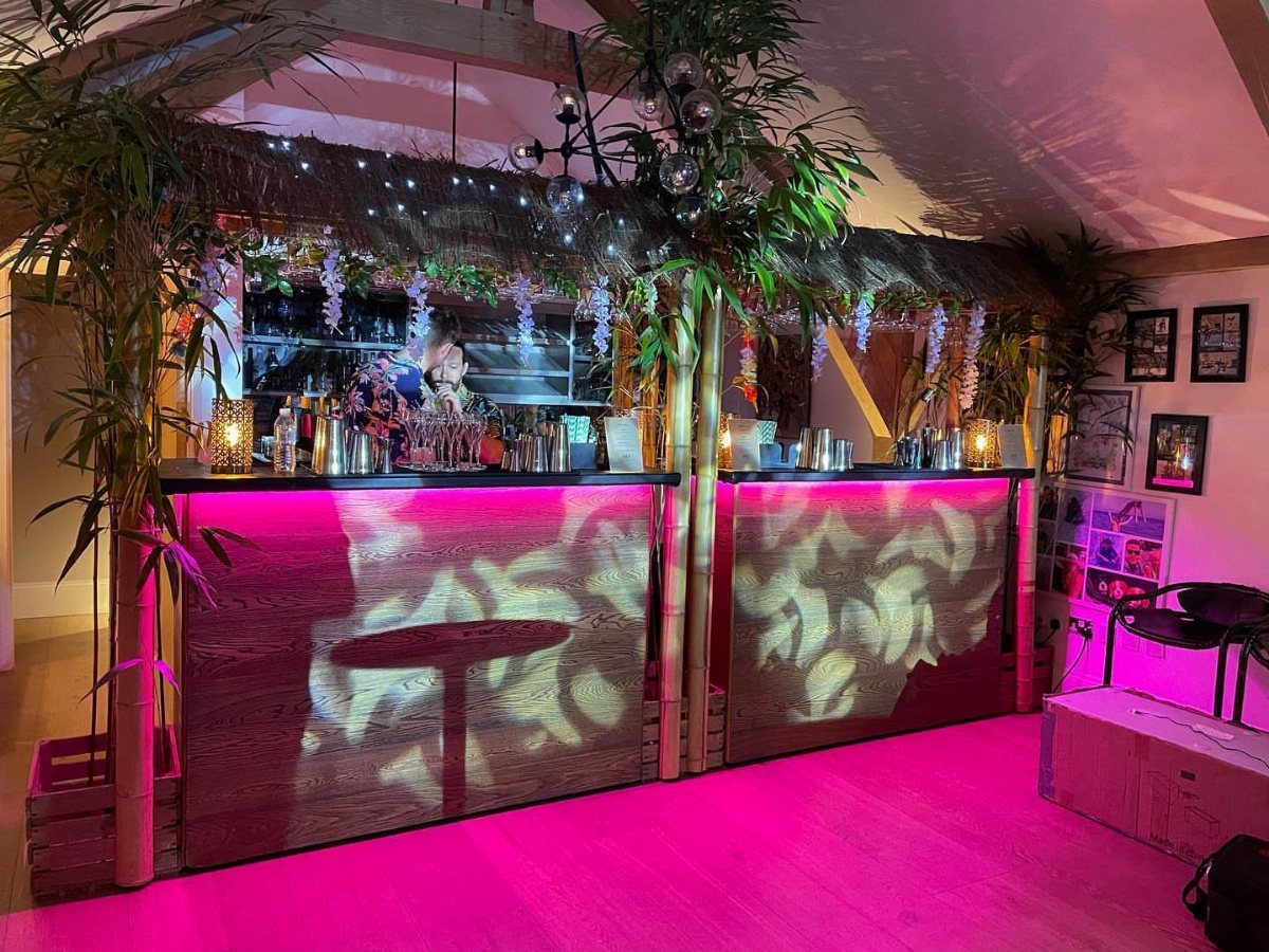 Tiki bar setup with thatched roof, bamboo, and pink neon lighting for a party.