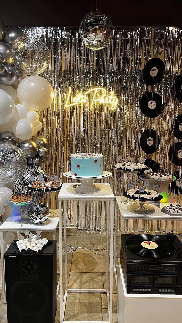 Party setup with silver tinsel, neon sign, blue cake, and disco balls.