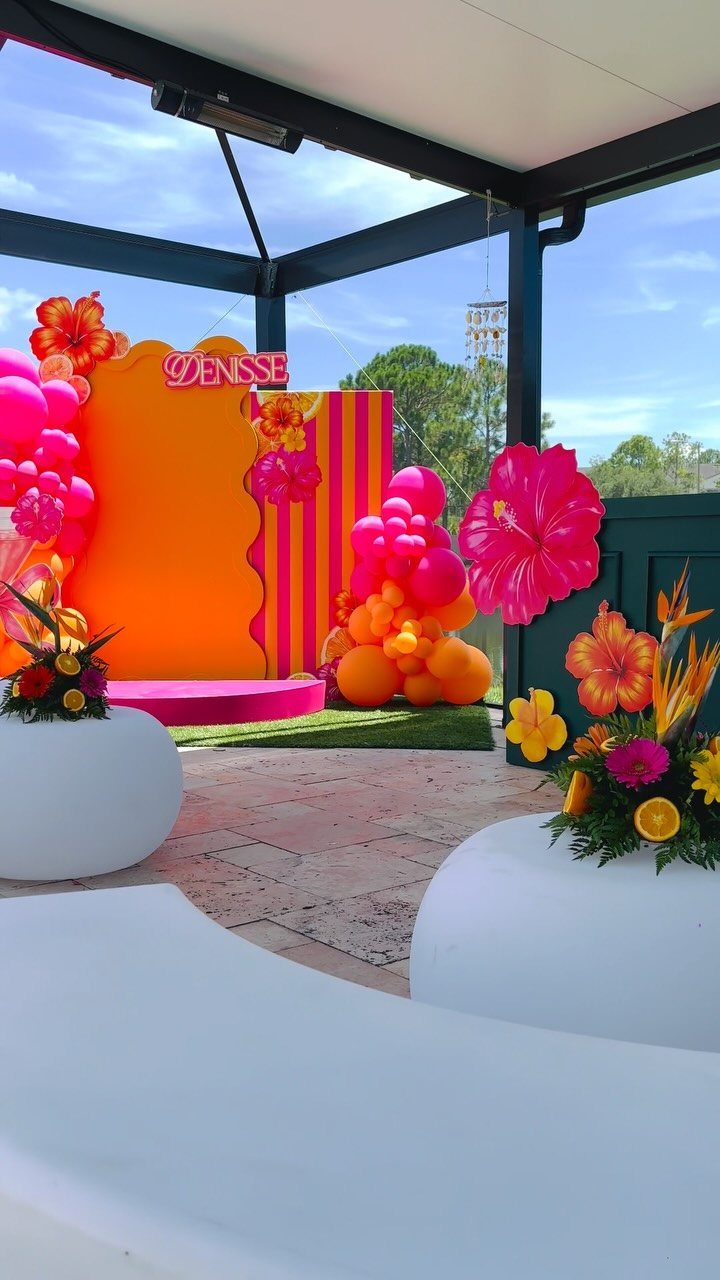 Outdoor party setup with orange backdrop, pink balloons, and large hibiscus flowers.
