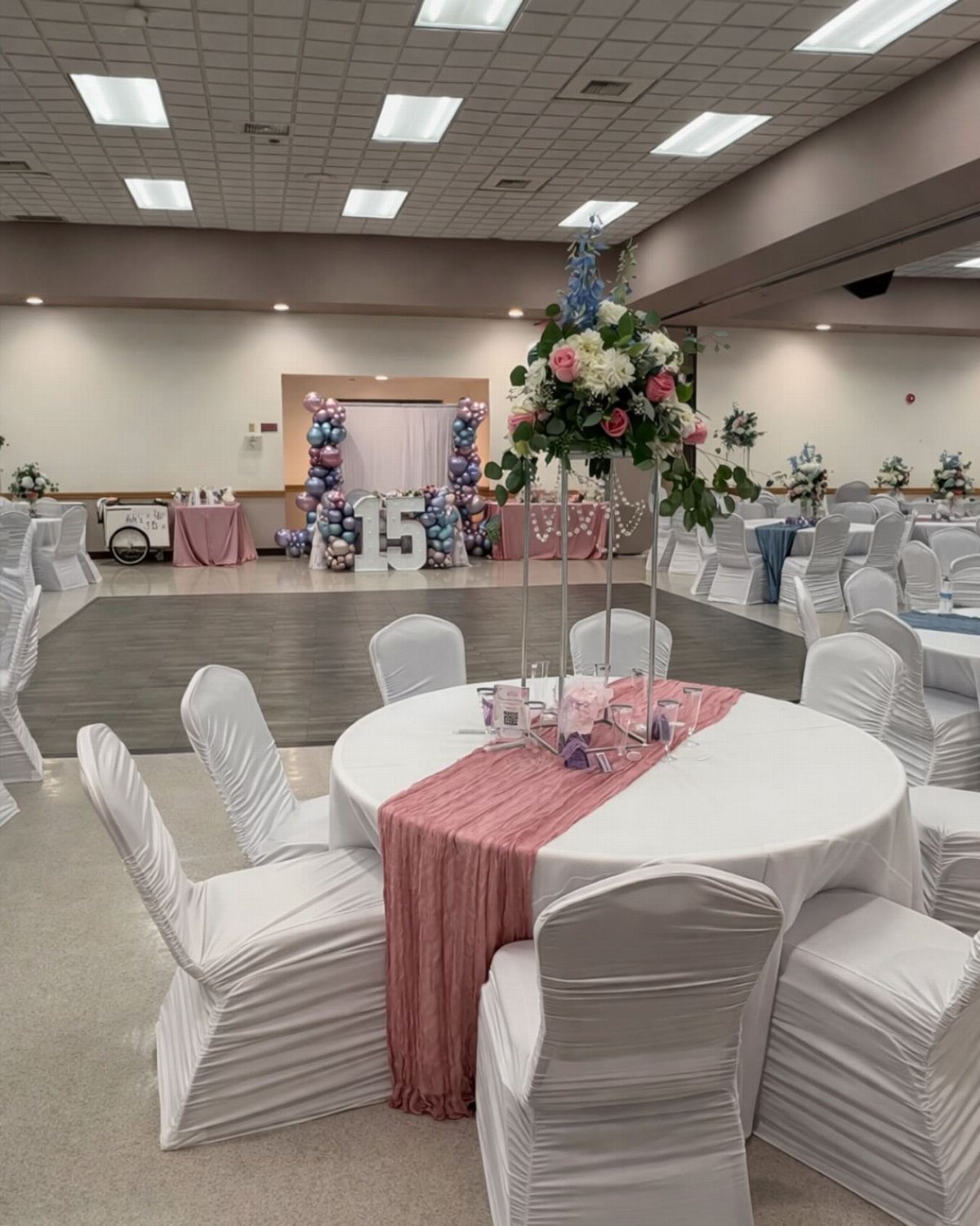 Decorated event hall with white tables, pink runners, and balloon arch for party.