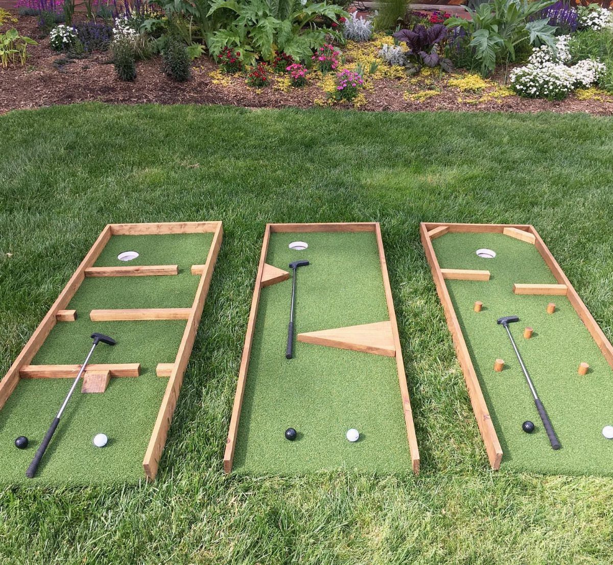 Three wooden miniature golf lanes with putters and balls on a green lawn.