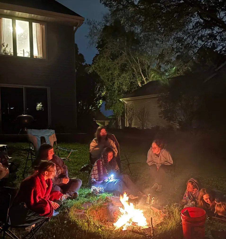People sitting around a glowing campfire in a backyard at night.