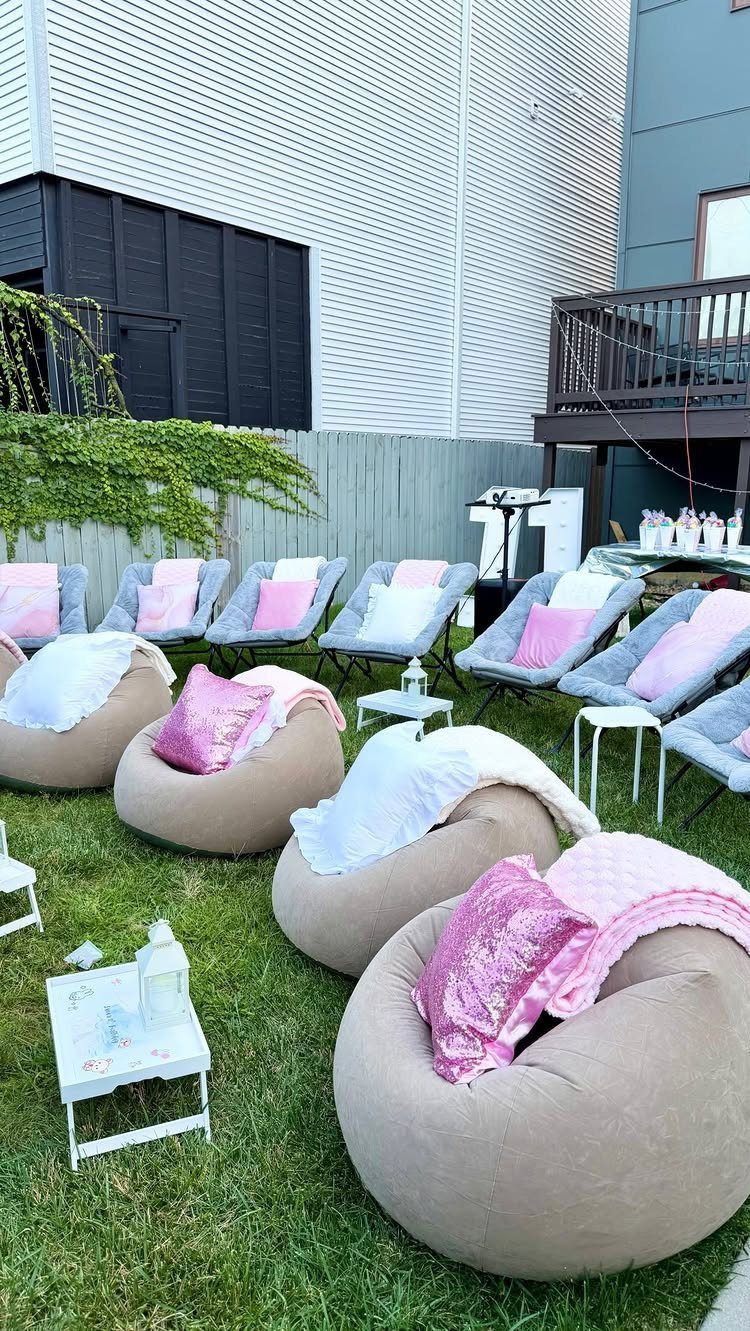 Outdoor lounge area with grey bean bags, pink pillows, and white blankets on grass.