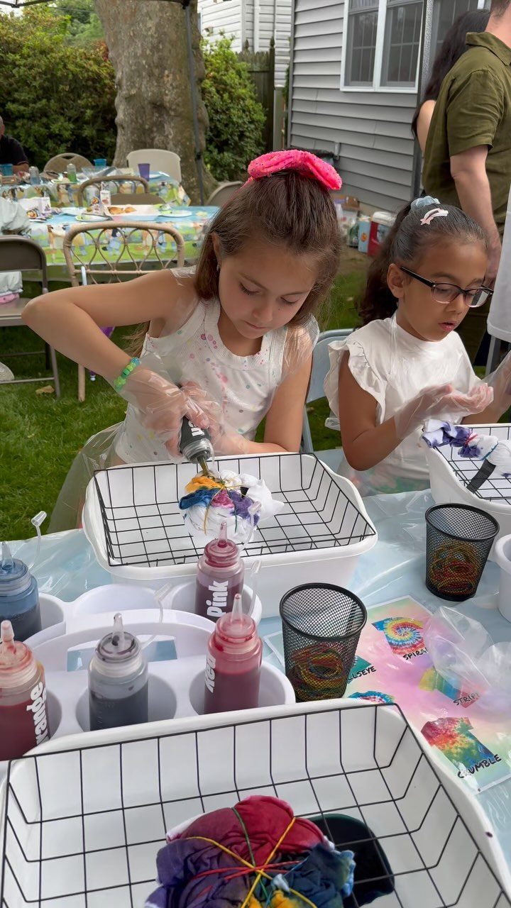 Two young girls tie-dyeing fabric at an outdoor table during a birthday party.