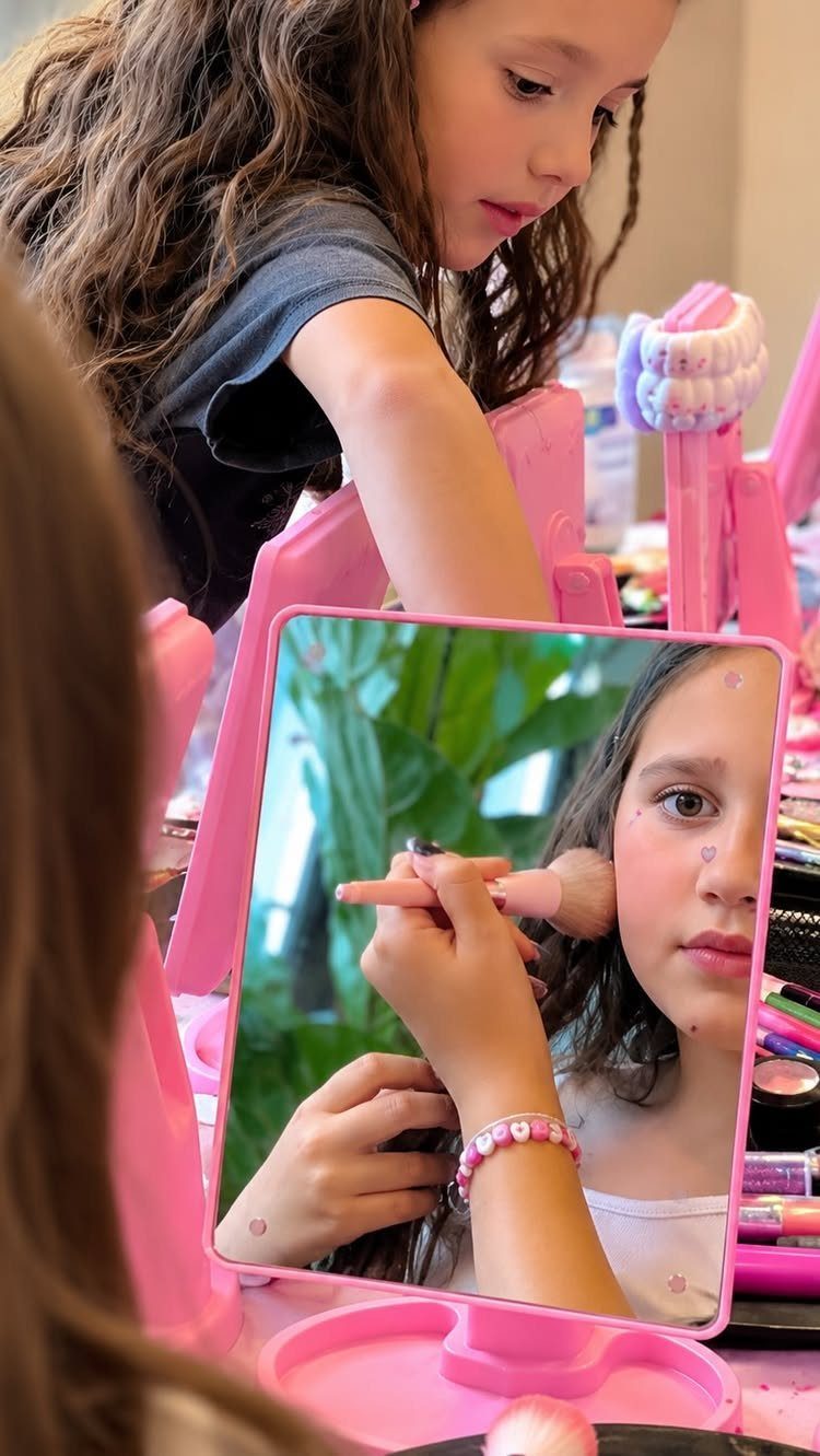 Young girl applying makeup with a brush while looking into a pink mirror
