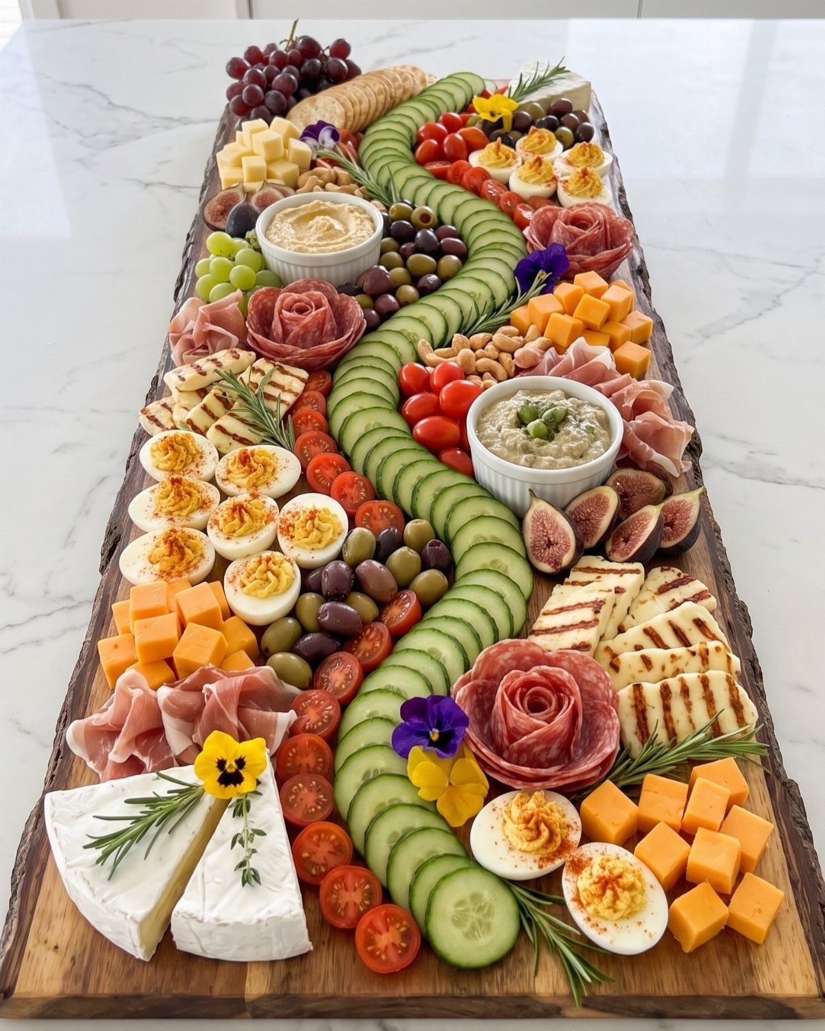 Charcuterie board with cucumbers, cheeses, meats, and dips on a white marble surface.