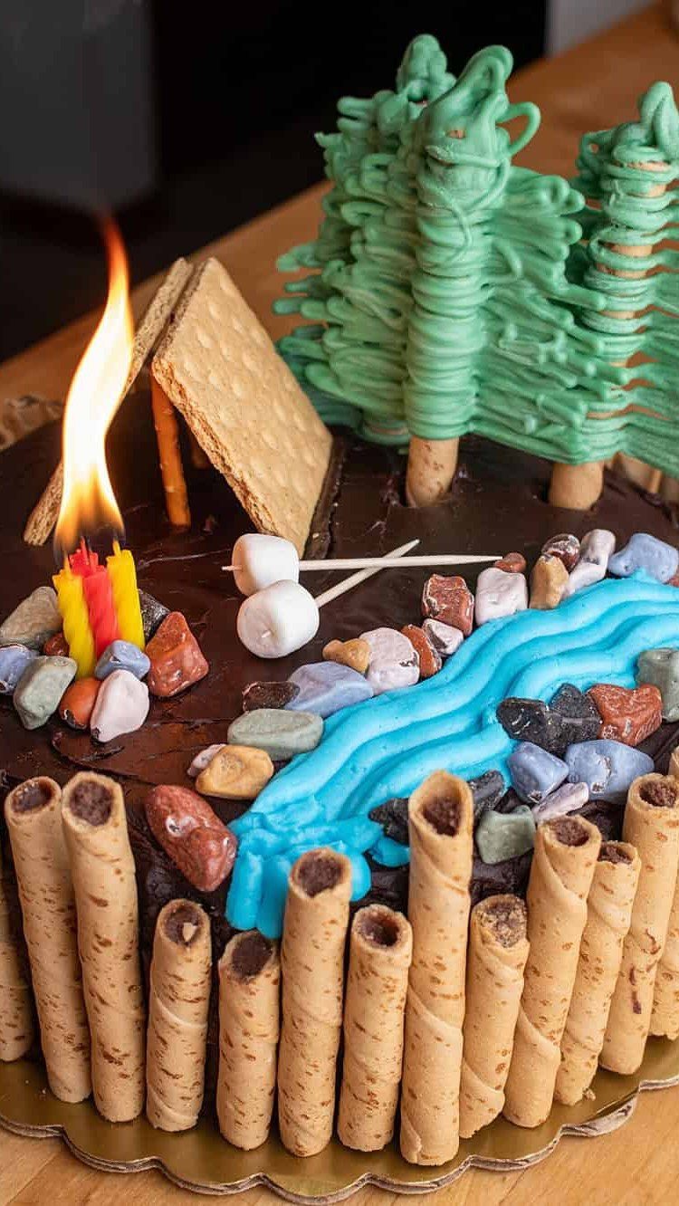 Camping-themed cake with chocolate frosting, wafer logs, graham cracker tents, and green trees.