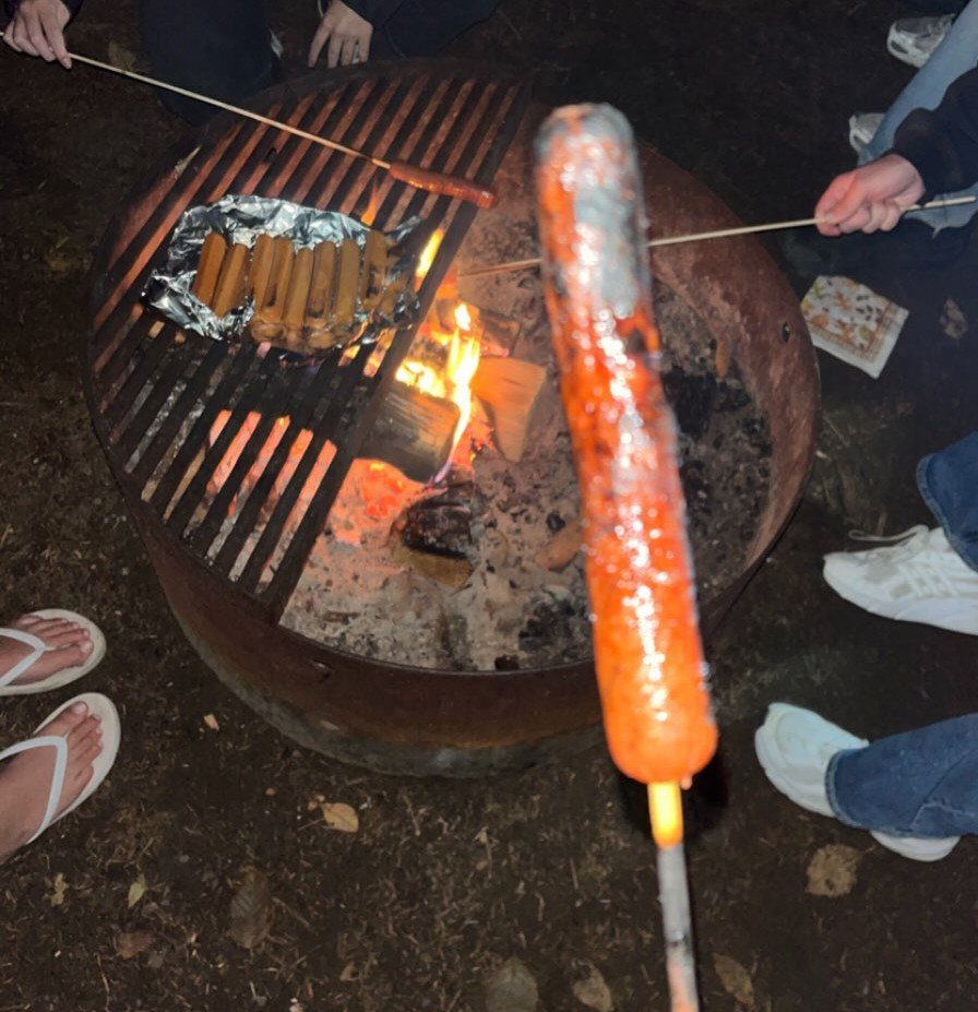 Hot dog on a stick roasting over a campfire in a metal pit.