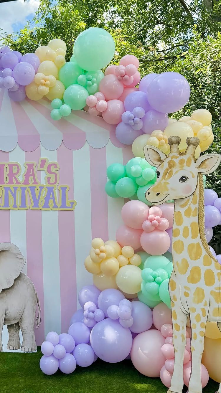 Pastel balloon arch framing a pink striped carnival backdrop with giraffe and elephant cutouts.