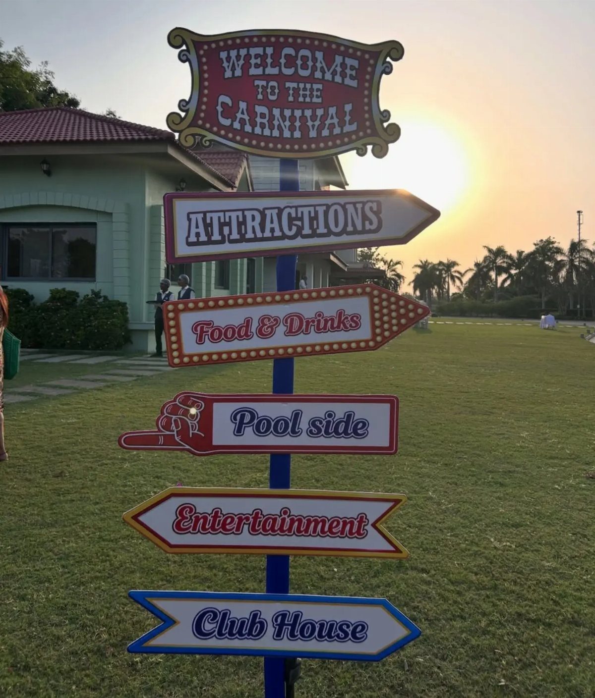 Carnival signpost with multiple directional arrows on a grassy lawn at sunset.