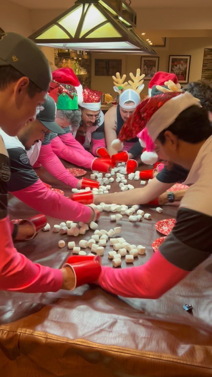 People in Santa hats using red cups to push marshmallows across a table.