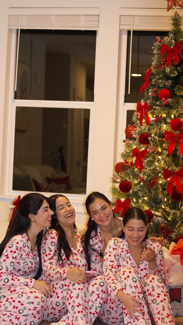 Four women in matching holiday pajamas laughing next to a decorated Christmas tree.