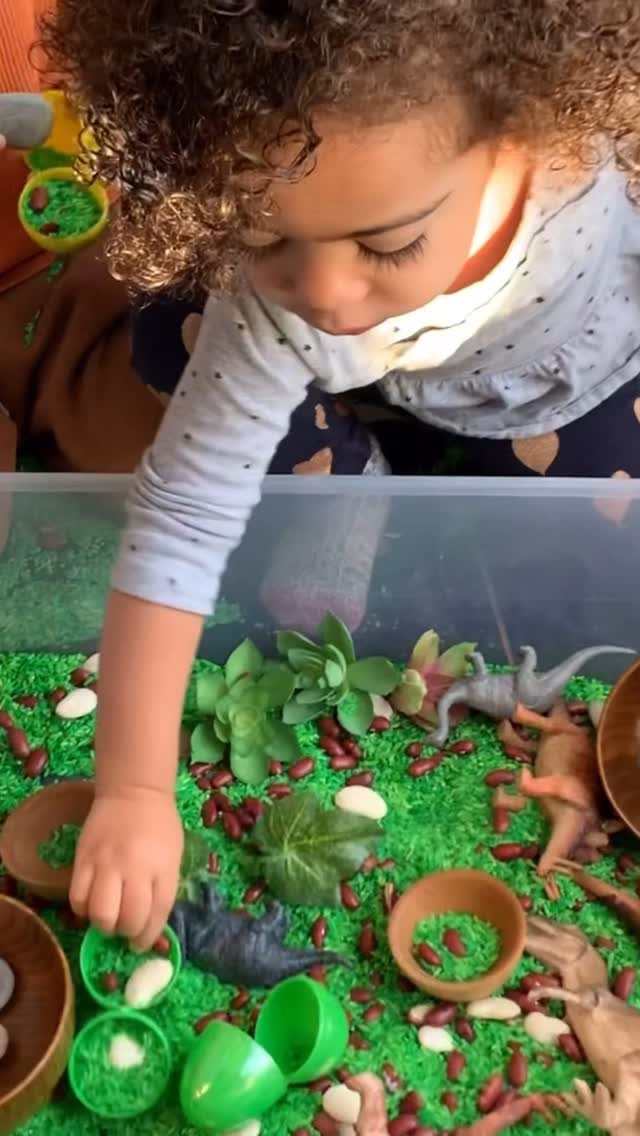 Child playing with plastic dinosaurs in a sensory bin with green grass and stones