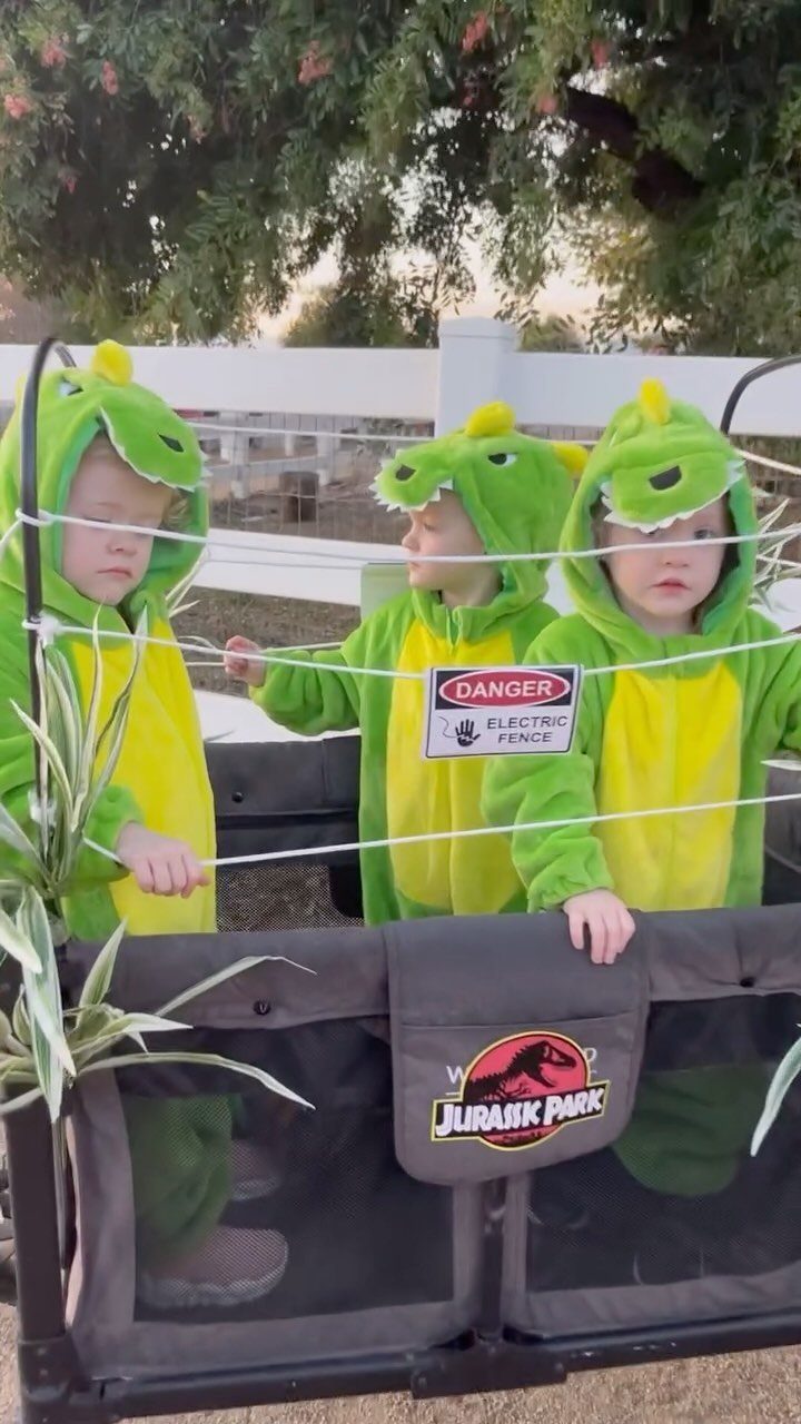 Three children in green dinosaur costumes standing behind a Jurassic Park themed fence.