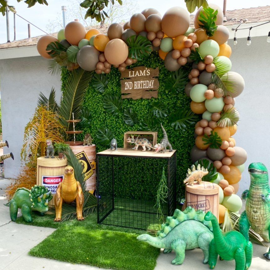 Dinosaur party decorations with a green foliage wall, balloons, and dinosaur figurines.