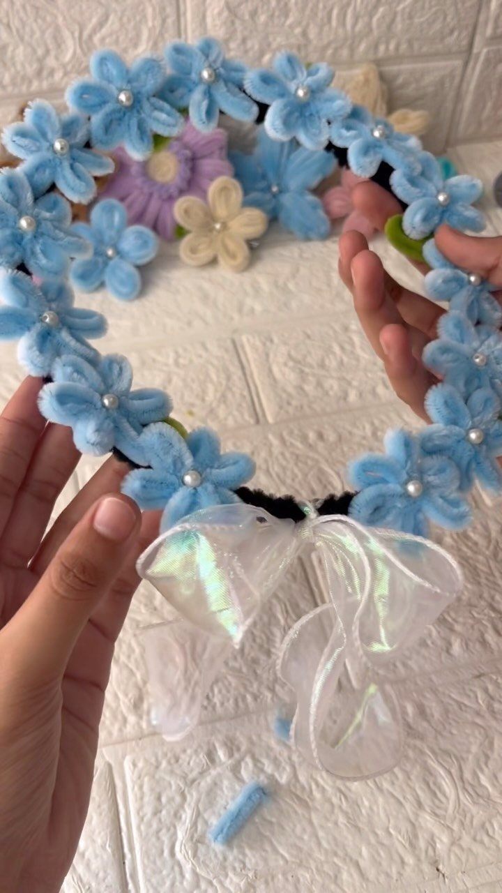 Light blue floral crown with pearl centers and an iridescent bow held by hands.