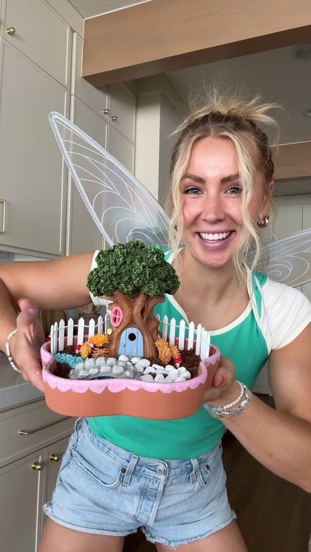 Woman wearing fairy wings holding a miniature DIY fairy garden with a tiny house.