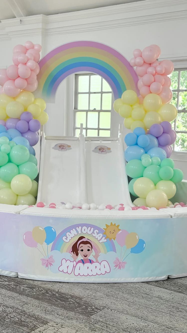White ball pit with slides, pastel balloon arches, and a rainbow backdrop.