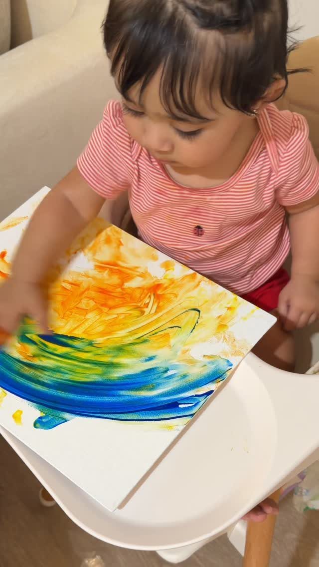 Toddler in striped shirt finger painting with blue and yellow paint on canvas.