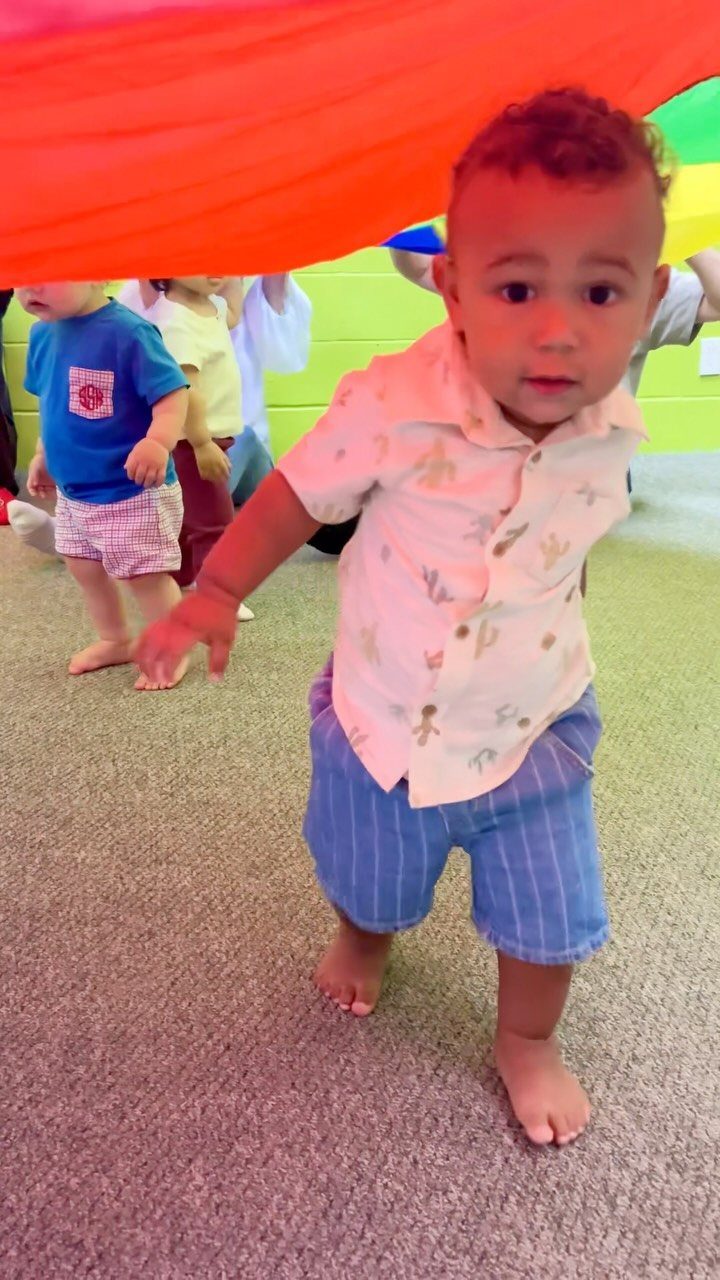 Toddler boy standing under a large orange parachute fabric in a playroom.