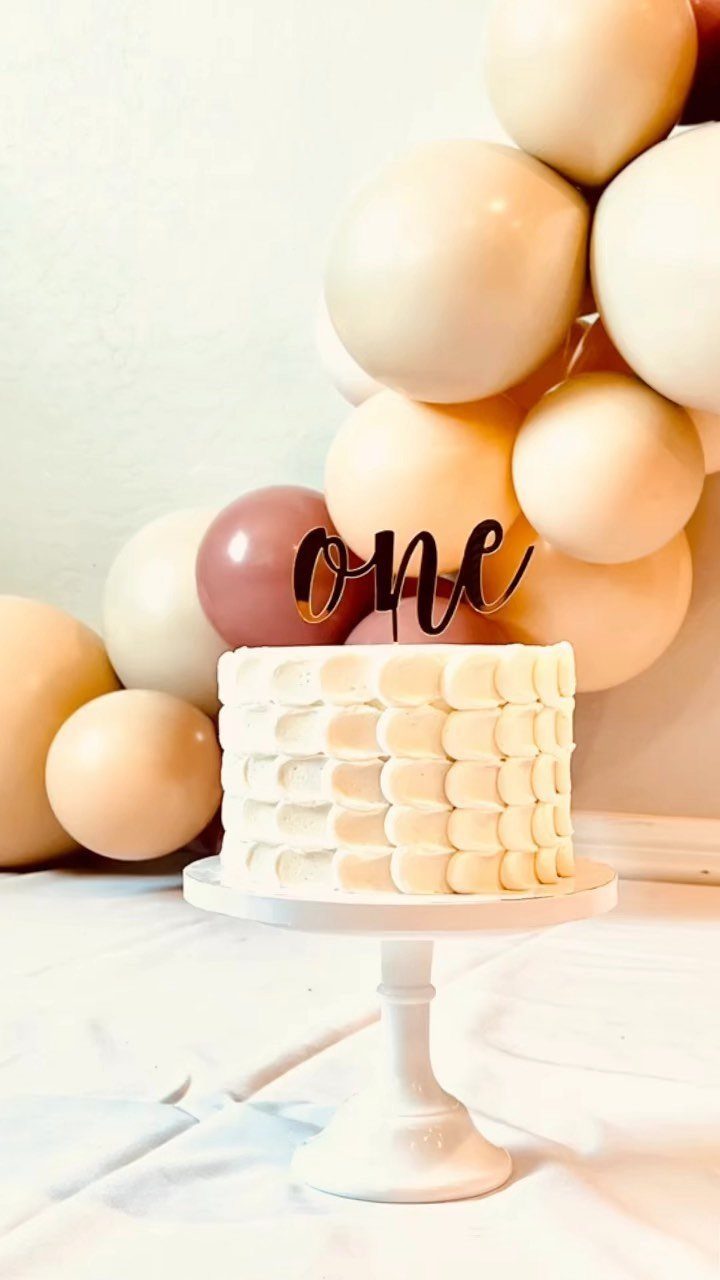 White frosted birthday cake with one topper on a stand with balloons behind.