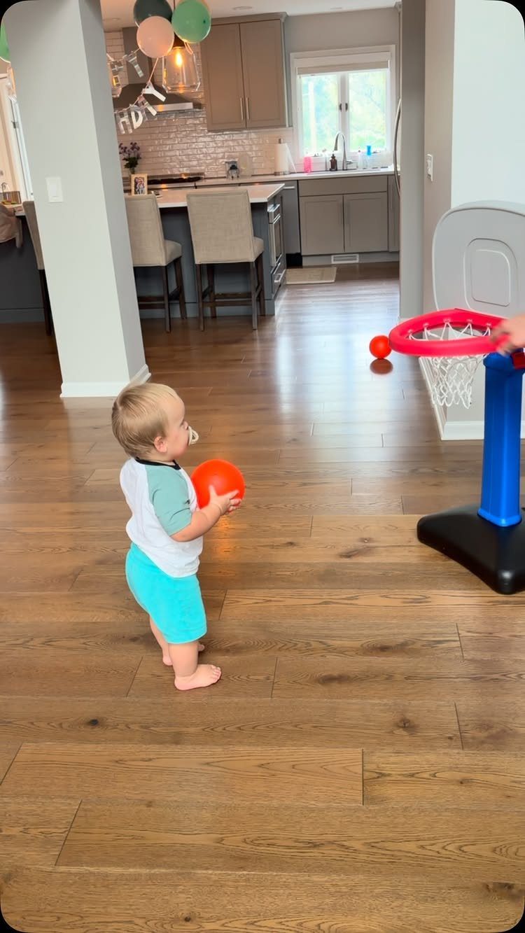 Toddler holding an orange ball in front of a toy basketball hoop indoors.