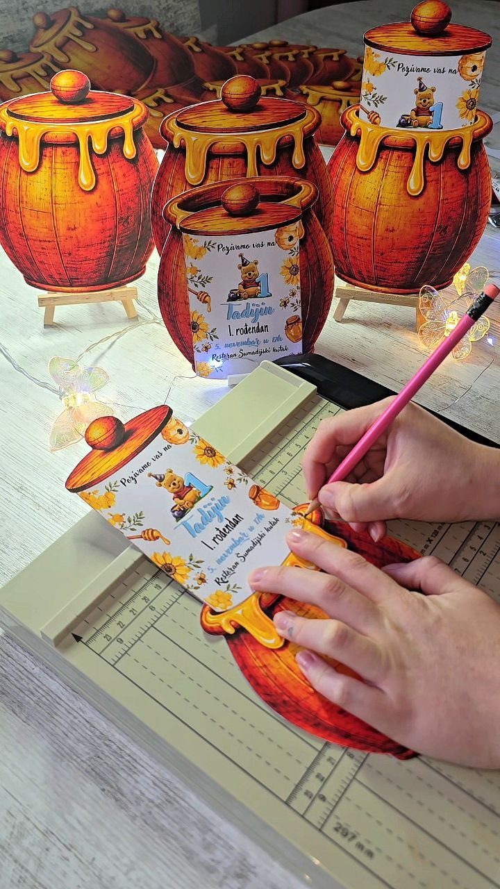 Hands assembling a 3D honey pot shaped first birthday invitation on a mat.