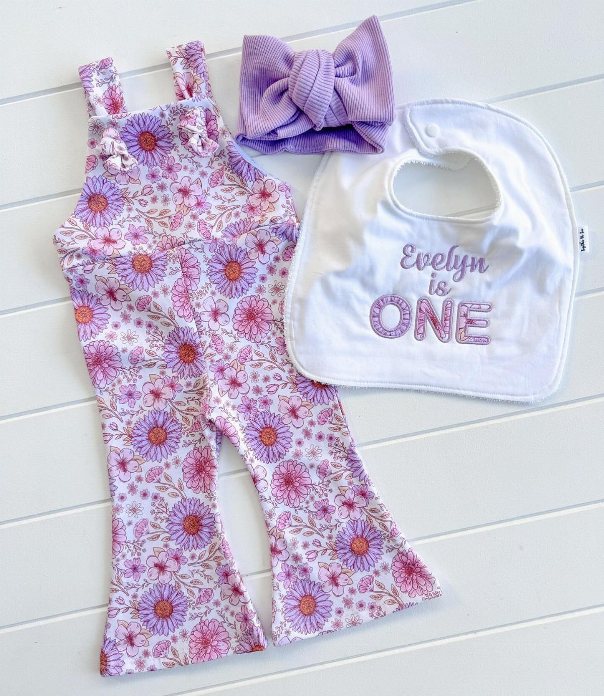 Floral baby overalls, purple headband, and a white birthday bib on white wood.