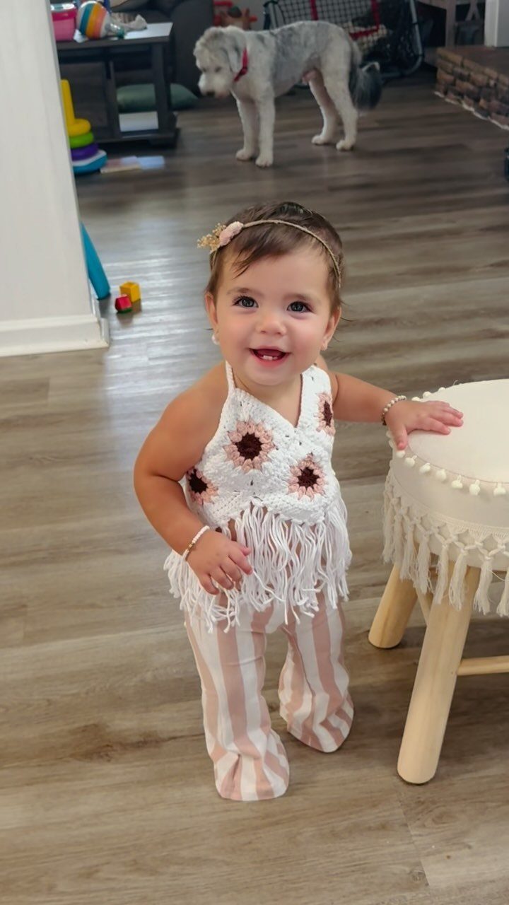 Baby girl wearing a crochet floral top and striped pants smiling indoors.