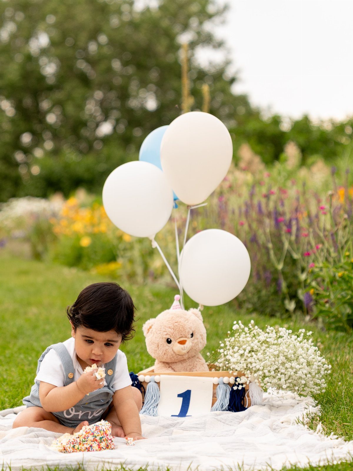 Baby in denim overalls sitting on a blanket with birthday balloons and cake.