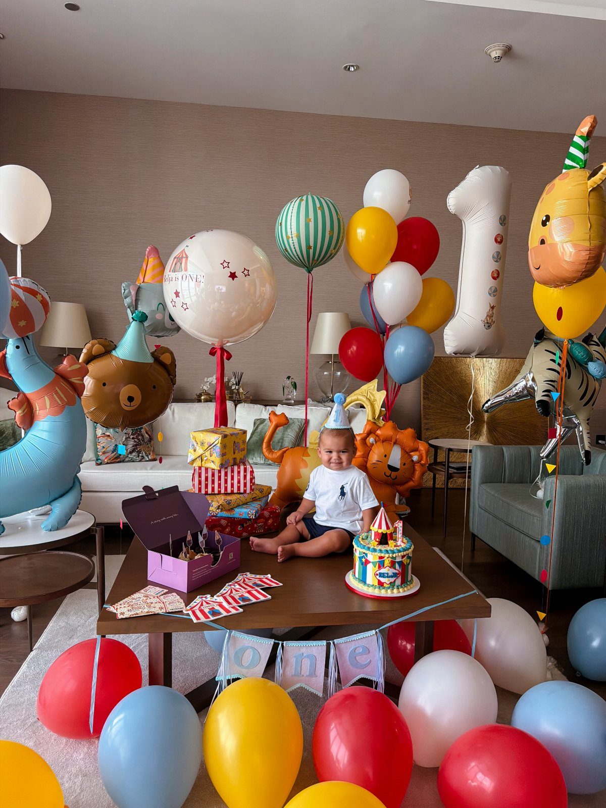 Baby in party hat sitting with colorful balloons and circus-themed decorations for first birthday.