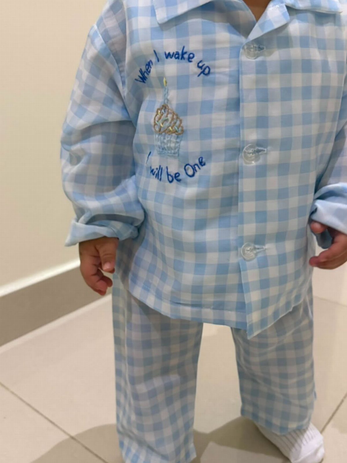 Toddler in blue gingham pajamas with an embroidered cupcake and first birthday text.