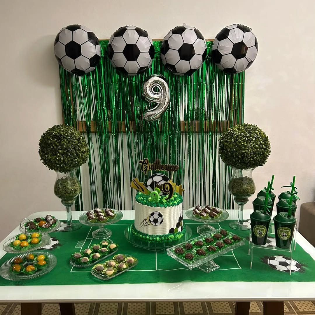 Football themed party table with soccer ball balloons, cake, and green snacks.
