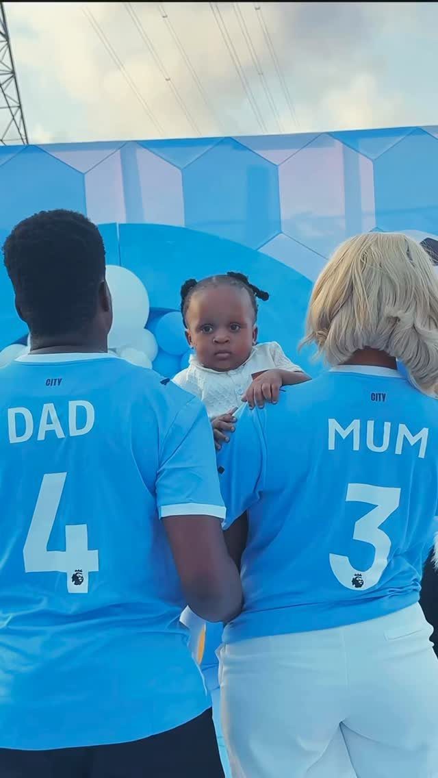 Parents in matching blue football jerseys holding a child at a themed party.